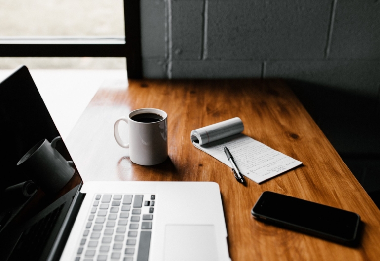 A polished wood desk, an open laptop, a phone, a notepad, a pen and a mug of black coffee.